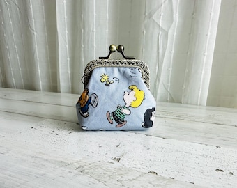May include: A light blue coin purse with a metal clasp and a cartoon print featuring Charlie Brown and Snoopy. The purse has a decorative border and is sitting on a white wooden surface.