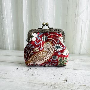 May include: A red fabric coin purse featuring a bird and floral design. The purse has a metal clasp and frame with a decorative pattern. The bird is brown and white, and the flowers are white and blue.