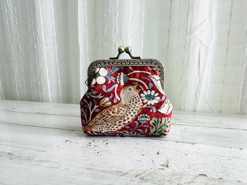 May include: A red fabric coin purse featuring a bird and floral design. The purse has a metal clasp and frame with a decorative pattern. The bird is brown and white, and the flowers are white and blue.