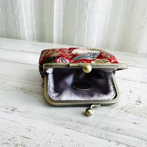 May include: Open, vintage-style coin purse with a red floral pattern. The purse has a metal frame with a ball clasp and a light purple satin lining. A coin rests inside the purse.