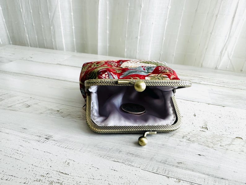 May include: Open, vintage-style coin purse with a red floral pattern. The purse has a metal frame with a ball clasp and a light purple satin lining. A coin rests inside the purse.