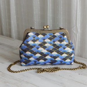 May include: A blue, white, and gold fan-patterned clutch purse with a brass clasp and chain strap. The purse is rectangular with a rounded top and a decorative clasp. The chain is a gold color.