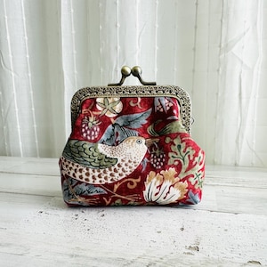 May include: A red fabric coin purse with a decorative metal clasp. The purse features a bird and floral pattern in shades of green, white, and gold. The background is a white wooden surface.