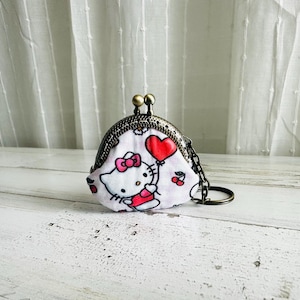 May include: A small, white coin purse with a metal clasp and chain. The purse features a cartoon character design with a red heart balloon and cherries. The background is a white and light wood surface.