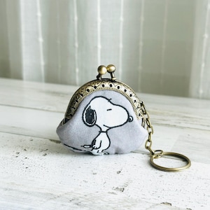 May include: A small, light gray fabric coin purse featuring a black and white illustration of the character Snoopy. The pouch has a vintage-style bronze metal kiss-lock clasp and an attached bronze metal chain with a circular key ring.