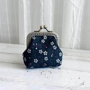 May include: A small coin purse made of dark navy blue fabric with a white cherry blossom pattern and subtle grid lines. The purse features an antique brass-toned metal kiss-lock clasp with an intricate filigree design along the top frame.