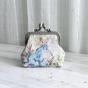May include: A small coin purse featuring a light-colored fabric with a printed illustration of a blue donkey character with a pink butterfly on its head. The purse has an ornate, antique-style bronze metal kiss-lock clasp frame at the top.