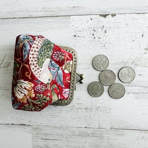 May include: A red patterned coin purse with a bird and floral design, and a metal clasp. Several coins are next to the purse on a white wooden surface. The purse has a vintage style.