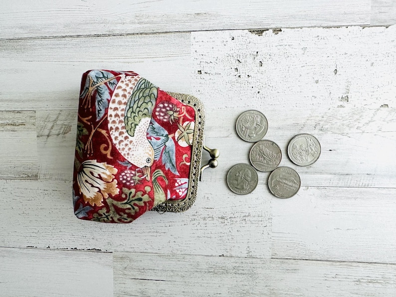 May include: A red patterned coin purse with a bird and floral design, and a metal clasp. Several coins are next to the purse on a white wooden surface. The purse has a vintage style.