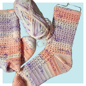 Puede incluir: Un par de calcetines tejidos a crochet en tonos rosa, morado y blanco. Los calcetines están hechos con un punto de encaje y tienen un hilo jaspeado con motas de color. También se puede ver una madeja de hilo en la imagen.