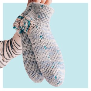 May include: A pair of crocheted socks in a light gray color with blue and pink stripes. The socks have a textured pattern and are made with a soft yarn.