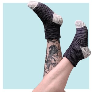 Op de afbeelding: Een paar zwarte en witte gehaakte sokken met een gestreept patroon. De sokken worden gedragen op de benen van een persoon met een zwarte en witte tatoeage.