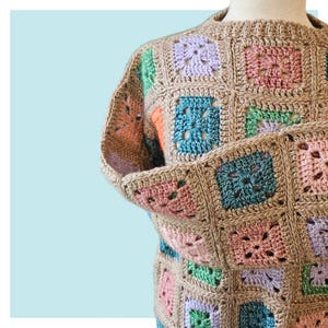 May include: A tan crochet sweater with a square pattern. The squares are in various pastel colors, including pink, blue, green, and purple. The sweater has long sleeves and a round neckline. The yarn appears to be a natural fiber.