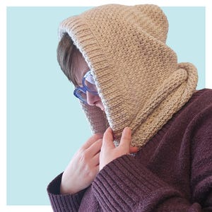 May include: A beige knitted hooded scarf with a textured pattern. The scarf is worn over a person's head and pulled up to cover their face. The person is wearing a maroon sweater.