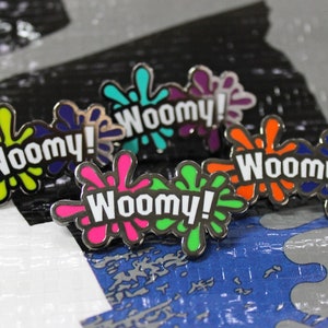 Peut inclure: Un ensemble de six épingles en émail avec un motif de projection coloré et le mot "Woomy!" en lettres blanches. Les épingles sont de différentes couleurs, notamment le vert, le jaune, le rose, le bleu, l'orange et le violet.