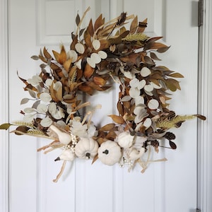 May include: A fall-themed wreath with dried leaves, white pumpkins, and sprigs of wheat. The wreath is a natural brown color with white accents.