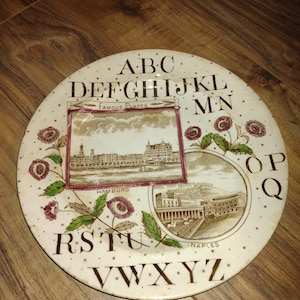 May include: A white ceramic plate with a brown alphabet border and two images of famous places, Hamburg and Naples. The plate has a brown and red floral design with gold stars.