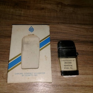 May include: A white box with a blue and gold stripe design. The box has a blue water drop logo and the text "CHROME COMPACT VU-LIGHTER BY SCRIPTO, INC." A black and silver lighter with the text "Compliments of JIM DODD Advertising Scripto, Inc." is next to the box.