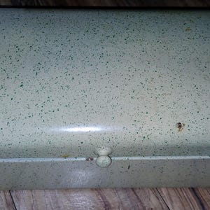 May include: A vintage, rectangular bread box with a speckled, light green finish. The metal box has a rounded top and a small, round knob on the front. There are signs of wear, including rust spots. The bread box is on a wooden surface.