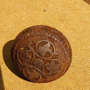 May include: A rusty metal button with a decorative floral design. The button has a raised edge and a smooth, rounded surface.