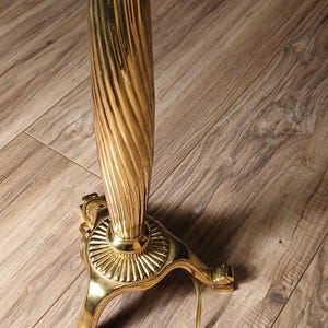 May include: A gold-colored floor lamp with a decorative, fluted column and ornate base. The lamp has a three-legged base and a yellow electrical cord with a two-prong plug. The lamp is on a wooden floor.