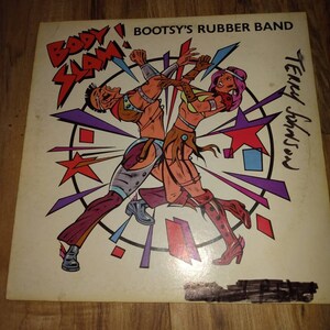 May include: A vintage vinyl record album cover featuring a colorful illustration of two figures in a wrestling match. The album is titled "Body Slam" by Bootsy's Rubber Band, with the text "Terry Johnson" written in the bottom right corner.
