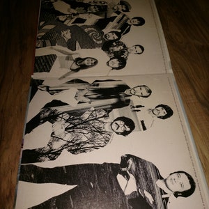 May include: A black and white photo of a band, the members are standing in a row, some are looking at the camera, others are looking away. The photo is on a white background with the band's name printed at the bottom.