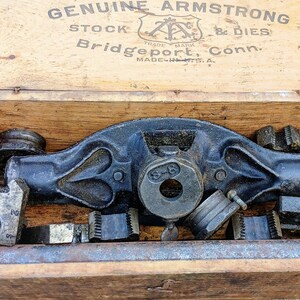 May include: A vintage wooden toolbox with the text "GENUINE ARMSTRONG STOCK & DIES" printed on the lid. Inside, a collection of metal tools, including a tap wrench and various dies, are neatly arranged. The tools are dark gray and show signs of wear.