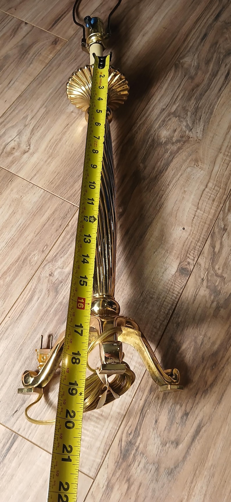 May include: A vintage brass lamp base, approximately 22 inches tall, with a fluted column and ornate base. The lamp has a decorative shell-like element near the top and a three-footed base. The lamp is unlit and has a cord.