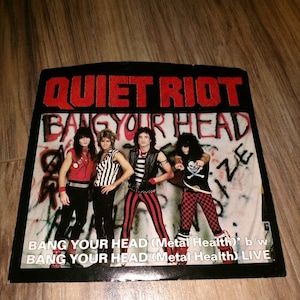 May include: A black and white vinyl record with a picture of the band Quiet Riot on the cover. The band members are wearing black and red striped shirts and black pants. The record title is "Bang Your Head (Metal Health)* b/w Bang Your Head (Metal Health) LIVE".