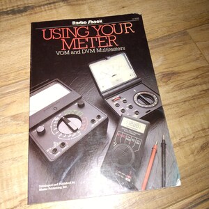May include: A book titled "Using Your Meter" by Radio Shack, featuring a black and silver analog multimeter and a black digital multimeter with red and black test leads.