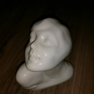 May include: White ceramic bust sculpture of a person's head and shoulders. The sculpture is smooth and has a glossy finish.