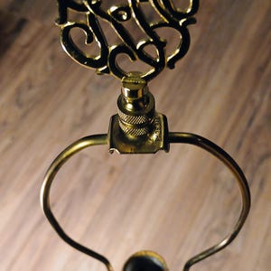 May include: Close-up of a gold-colored lamp harp with an ornate, swirling design at the top. The lamp harp has a curved handle and a threaded base for attaching to a lamp. The lamp base is also gold-colored.