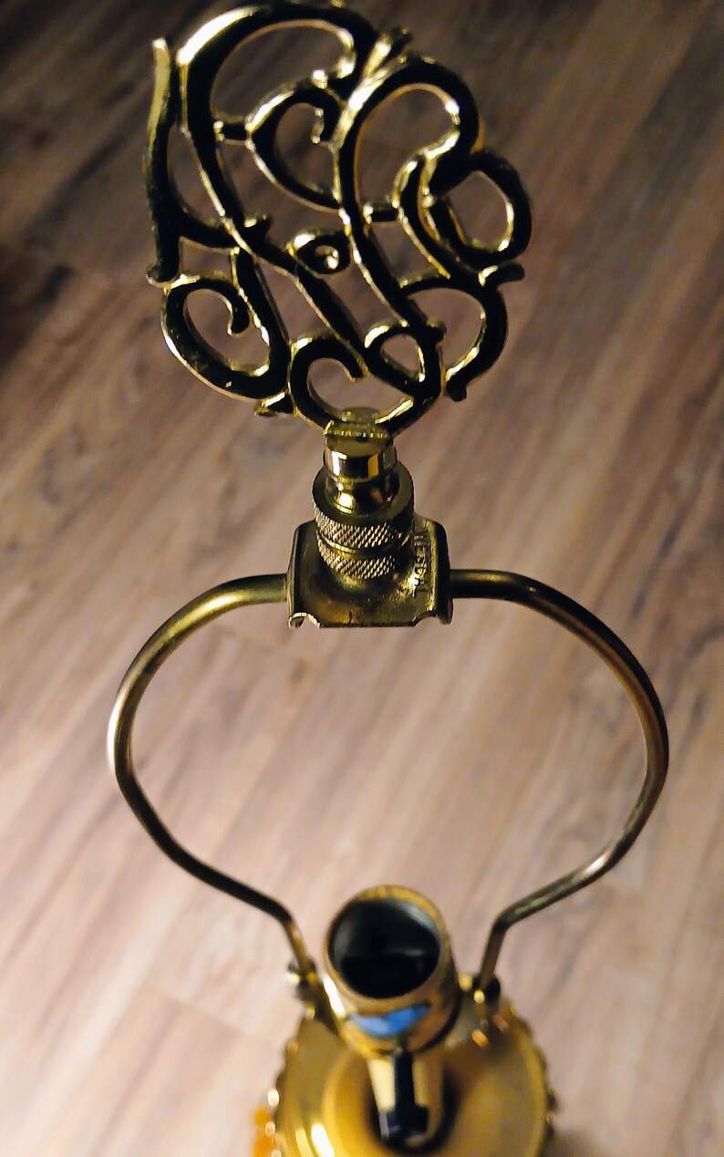 May include: Close-up of a gold-colored lamp harp with an ornate, swirling design at the top. The lamp harp has a curved handle and a threaded base for attaching to a lamp. The lamp base is also gold-colored.