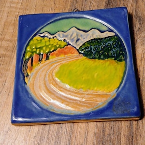Puede incluir: Azulejo cerámico cuadrado con una escena de paisaje. El azulejo presenta un camino sinuoso, campos verdes, árboles y montañas, todo dentro de un borde azul. La obra de arte utiliza tonos de verde, amarillo, naranja y azul.