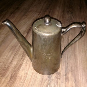May include: A silver teapot with a round lid and a handle. The teapot has a spout and a small inscription on the side that reads "PLATED DE  METZ".