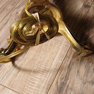 May include: A gold-colored Christmas tree stand with a decorative design. The stand has three legs and a central support column. A bundle of gold-colored wire is tied to the stand with a cream-colored ribbon.