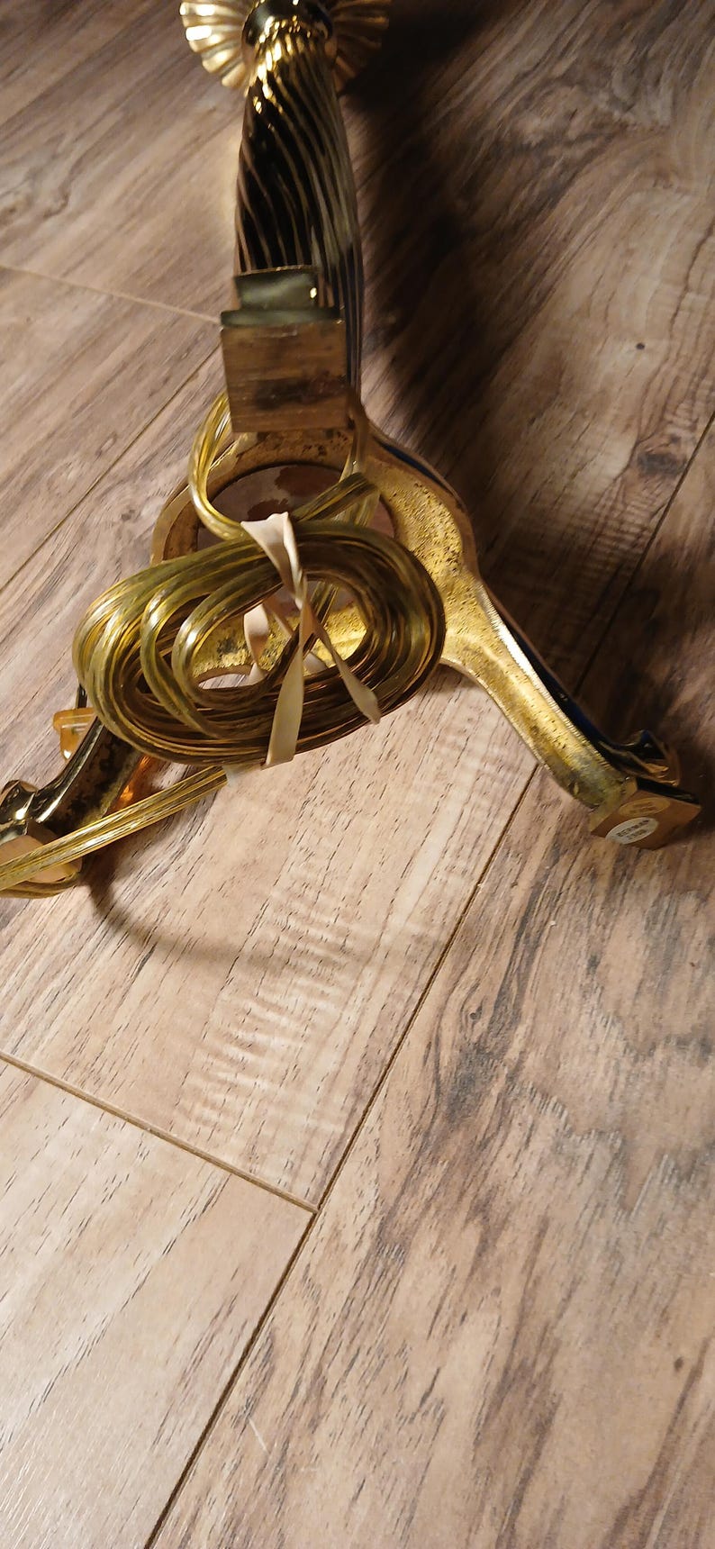 May include: A gold-colored Christmas tree stand with a decorative design. The stand has three legs and a central support column. A bundle of gold-colored wire is tied to the stand with a cream-colored ribbon.