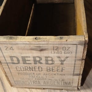 May include: Wooden box with the text "DERBY CORNED BEEF" and "12 OZ (340 GR)". The box is made of light-colored wood and has a rectangular shape. The text is printed in a dark font.