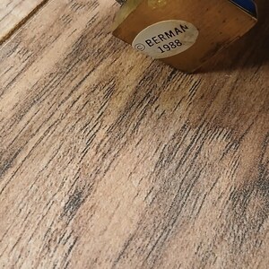 May include: A vintage brass decorative object with a blue and gold accent. The base is wooden, and a sticker reads "BERMAN 1988". The object has a curved, ornate design and is set against a wooden surface.