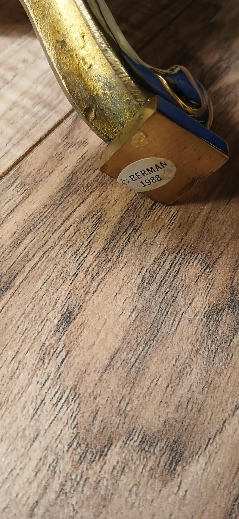 May include: A vintage brass decorative object with a blue and gold accent. The base is wooden, and a sticker reads "BERMAN 1988". The object has a curved, ornate design and is set against a wooden surface.