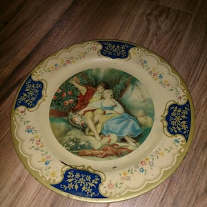 May include: A vintage tin plate with a blue and gold border and a floral design. The center of the plate features a painting of a couple in a garden setting.