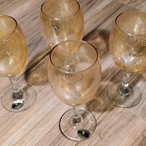 May include: Four amber-colored wine glasses with clear stems and bases. Each glass has a decorative etched pattern. The glasses are arranged on a wooden surface.