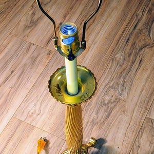 May include: A vintage brass table lamp with a decorative floral finial. The lamp features a fluted column, a scalloped base, and a curved arm holding the light bulb. The lamp is on a wooden floor.