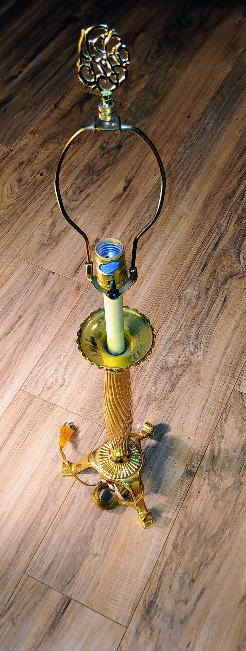May include: A vintage brass table lamp with a decorative floral finial. The lamp features a fluted column, a scalloped base, and a curved arm holding the light bulb. The lamp is on a wooden floor.