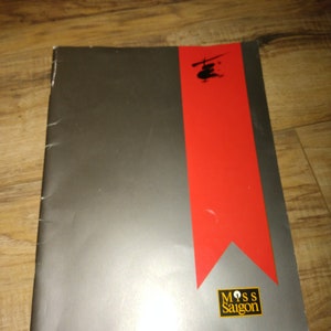 May include: A silver and red program for the musical Miss Saigon. The cover features a black silhouette of a helicopter.