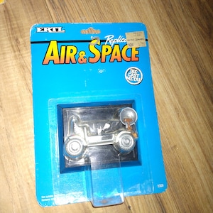 May include: A blue and white cardboard box with a silver die-cast toy vehicle inside. The box has the words "ERTL Replica Air & Space" printed on it. The toy vehicle is a silver lunar rover with a satellite dish on top. The box also has a sticker that says "Die Cast Metal".