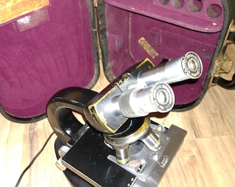May include: A vintage black microscope with a silver stage and two eyepieces. The microscope is in a black carrying case with a purple velvet lining.