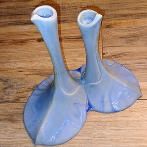 May include: A pair of light blue ceramic vases with a unique, organic shape. The vases have long, slender necks and flared bases, connected at the bottom. The color is a soft, pale blue with subtle variations.