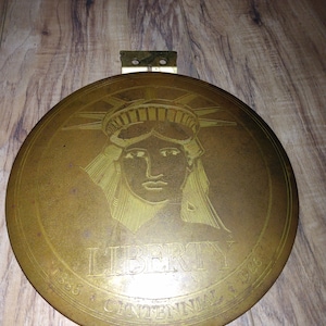 May include: A gold-colored round clock pendulum with a raised image of the Statue of Liberty and the text "LIBERTY CENTENNIAL 1886 - 1986".